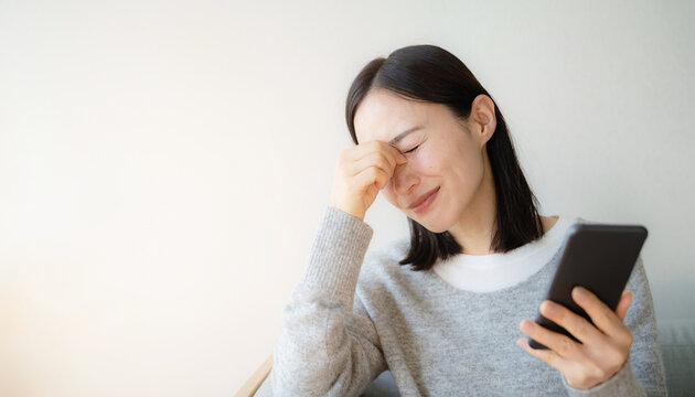 Stressed Japanese woman holding phone and touching her tired eyes スマホを持ちながら疲れた目を閉じて額を押さえる日本の女性