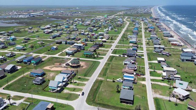 Surfside coastal span homes near Seashell Dr extend toward Gulf shoreline with bridge marsh corridor visible beyond neighborhood grid. Varied roof colors, grassy parcels, soft surf, Freeport, TX
