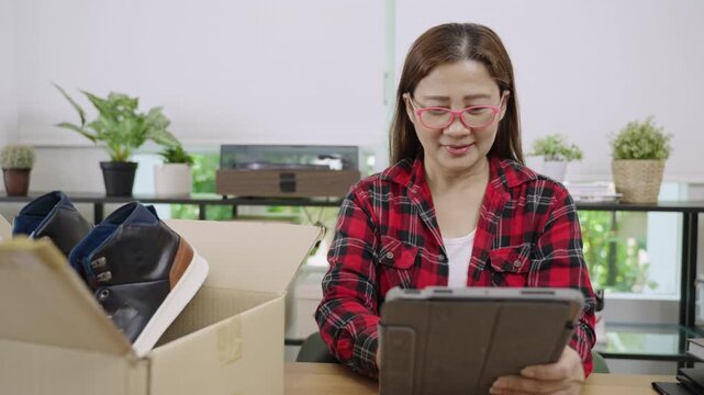 Asian woman entrepreneur, small business owner, with eyeglasses, using a digital tablet to check list of delivery from online shopping for customer in home office. E-commerce and SME business concept.