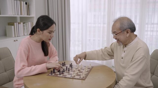 Happy beautiful young Asian woman sitting smiling and playing a chess game with her senior man father together having cheerful and enjoyable time during relaxation and leisure in living room at home. 