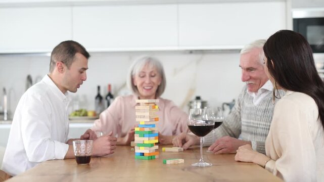 Joyful senior parents and young couple sitting around table playing brick game and drinking wine in goblets at home