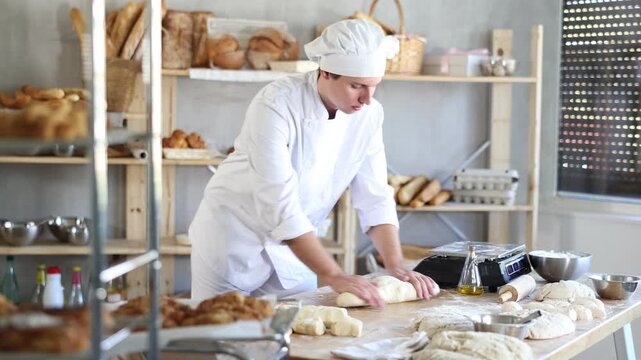 Young male baker prepares raw yeast dough in bakery, shaping it into a baguette and rolls dough to give the shape of future bread. High quality 4k footage