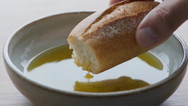 Touching golden oil, adult hand dipping torn bread into bowl at kitchen table, soaking for eating