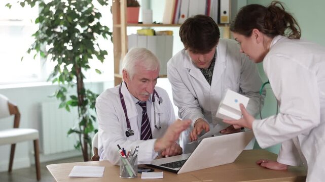 Three doctors bent over laptop. Mentor helps to trainee, teaches how to work with medical statistics program. Complex clinical case, medical consultation, symposium, meeting. 