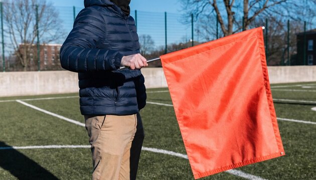 High resolution 8k photorealistic image of an anonymous football coach standing on the field holding a large blank colored flag, ready for custom text or team branding during a game day event.
