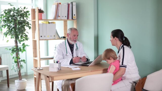 At clinic, woman with child at senior male doctors appointment. Medical specialist and patient sit at table and communicate, interview, anamnesis, patient complaints