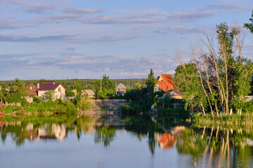 Fototapeta premium Picturesque countryside houses on river bank with bright reflections in calm water at sunset.