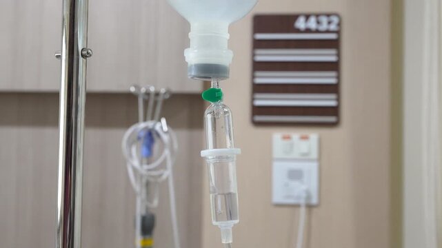 	
A close-up shot of an intravenous (IV) drip infusion set with saline solution drops in a hospital room, featuring a blurred background.