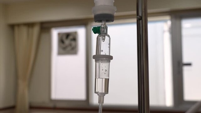	
A close-up shot of an intravenous (IV) drip infusion set with saline solution drops in a hospital room, featuring a blurred background.