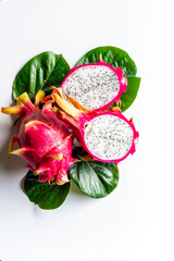 Obraz premium Two pitaya fruits - whole and cut in half - with green leaves on white table. Ripe dragon fruit or pitahaya. Tropical fruits, top view
