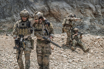 Obraz premium Military special forces soldier helping injured comrade walk during a combat mission in rocky terrain. Team members providing cover fire in background. Concept of brotherhood and rescue.