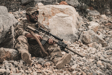 Obraz premium Exhausted military commando resting on rocky terrain during a combat mission. Tired armed forces operator catching his breath after battle. Tactical survival and warfare concept.