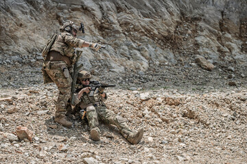 Obraz premium Two military special forces soldiers in combat action aiming weapons in a rugged rocky terrain. One soldier provides cover fire with a pistol while another sits aiming an assault rifle.