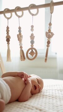 Curious newborn baby boy looking at hanging wooden toys while lying on a bed. Infant playing with a baby gym, exploring educational montessori toys in a bright, sunlit nursery