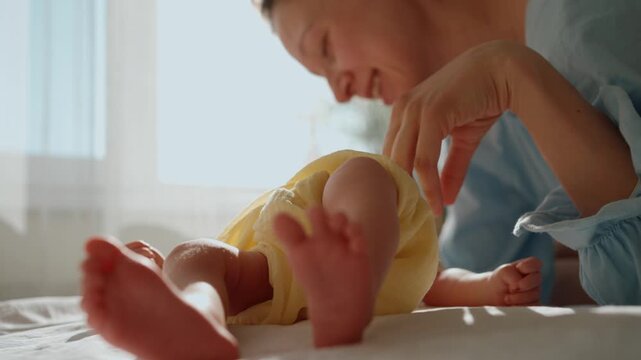 Loving young mother smiles and gently plays with her infant's tiny feet as they lie on a bed in a bright, sun drenched room, sharing a heartwarming and joyful connection