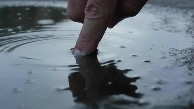 Hovering finger touching puddle on wet ground, making ripples, warping reflection, exploring water