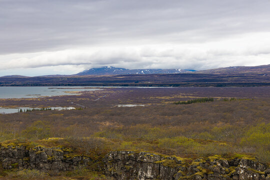 Vista panor&aacute;mica del Parque Nacional Thingvellir en Islandia bajo un cielo nublado, mostrando rocas de lava con musgo, llanuras de oto&ntilde;o, el lago Thingvallavatn y monta&ntilde;as nevadas lejanas.