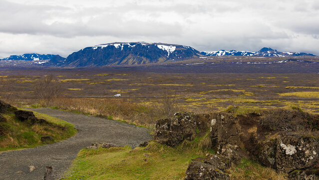 Un sendero de tierra serpentea por el Parque Nacional Thingvellir en Islandia, ofreciendo una vista panor&aacute;mica de extensas llanuras de tundra y una furgoneta blanca frente a monta&ntilde;as nevadas.