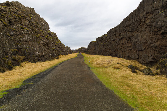 Un amplio sendero de tierra cruza el desfiladero de Almannagj&aacute; en el Parque Nacional Thingvellir, Islandia, flanqueado por enormes paredes de roca volc&aacute;nica y llanuras de hierba amarillenta. 