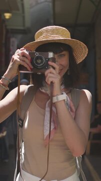 Vertical video. A woman in a wide straw hat holds a vintage camera to her face and looks through the viewfinder, as if capturing the moment. Warm, soft tones create a relaxed atmosphere.