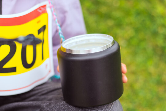 Individual holding a black insulated tumbler with a silver lid while wearing a race bib numbered 207, seated on grass in an outdoor setting