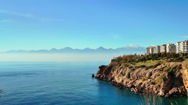 Misty taurus mountains rise behind blue mediterranean sea cliffs and antalya city skyline buildings.