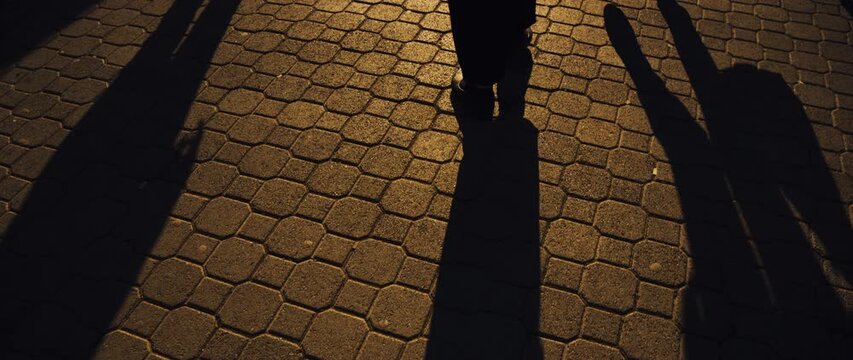 Long elongated shadows of pedestrians stretch across textured paving stones in warm golden-hour light, evoking mood, movement, urban life and quiet evening atmosphere, 4K.