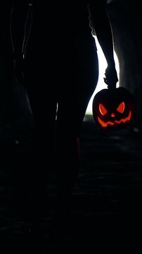 Vertical video. Silhouette Walking Through Dark Tunnel Carrying Glowing Jack O Lantern Toward Bright Light at Tunnel End, Halloween Mystery, Fear And Unknown Journey