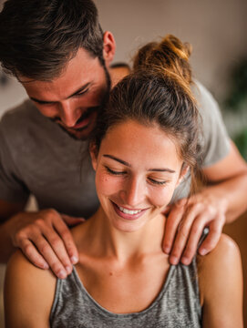 Young couple sharing a tender moment as man gently massages woman?s shoulders with warm smiles in a cozy indoor setting, expressing affection and care