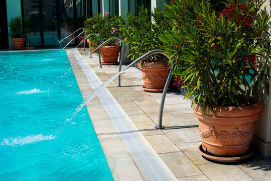 Outdoor hotel pool with water jets shooting across the pool surface, large terracotta pots with red and pink oleander plants placed along the pool edge beside metal handrails, tiled terrace and hotel 