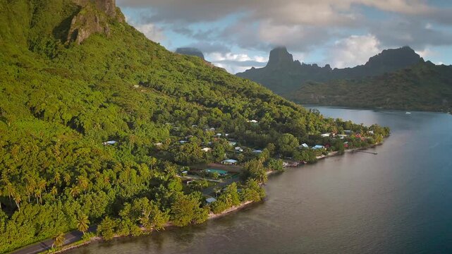 Moorea island vibrant coastal village nestled at the base of dramatic green mountains and dense tropical jungle, with clear South Pacific waters extending into the bay. Aerial view drone flight