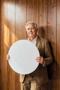 Senior man holds a large white circular sign in front of his torso against vertical wood paneling. Warm side lighting highlights the texture of his tan suit and gray hair.