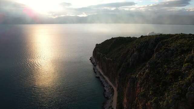 Aerial footage flying along the famous Arvanitia promenade path in Nafplio, Greece. Stone walkway carved between massive rocky cliffs andv splashing waves of the blue sea, golden sunset with sun path.