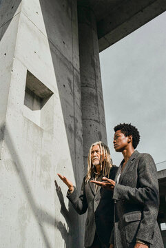 Man and woman stand beside a massive concrete pillar under an overcast sky. They gesture toward the textured grey structure while sunlight creates sharp shadow lines on the wall.