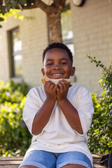 African American child sitting on bench in front yard holding sandwich in white tee, denim shorts © wavebreak3