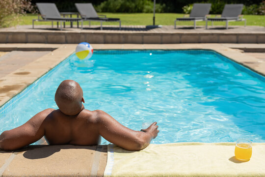 African American man reclining bare-chested at backyard pool edge, with glass, towel and beach ball