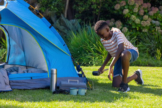 African-American teen kneeling, driving tent stake with mallet in garden in striped tee and shorts