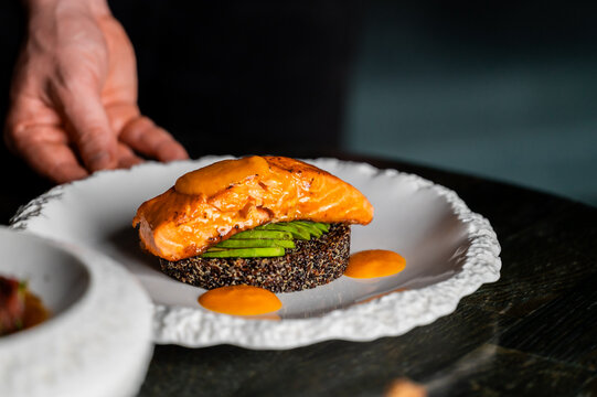 Seared salmon fillet served on a bed of dark quinoa and sliced avocado, plated beautifully with a vibrant orange sauce in a restaurant setting.