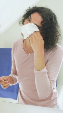 Vertical video: Leaning nonbinary person wiping face at bathroom mirror, cleansing with white cloth