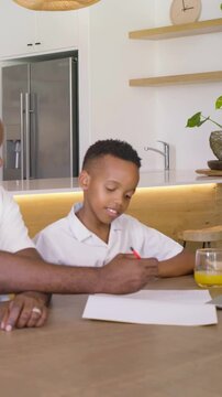 Vertical video: Father placing hand on son, guiding red pencil at kitchen table, teaching homework