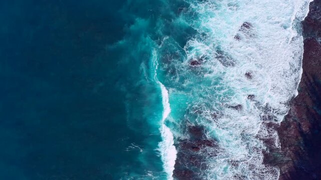 Aerial drone view of powerful ocean waves crashing against a rugged rocky coastline in Hawaii. Turquoise Pacific waters and swirling white sea foam create stunning natural patterns along the volcanic 