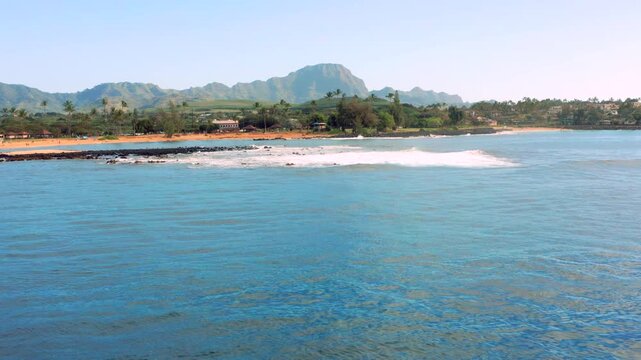 A beautiful, wide shot of a Hawaiian bay featuring vibrant blue and turquoise waters. Waves gently break over a dark rock jetty/breakwater near a golden sand beach. In the background, lush green tropi