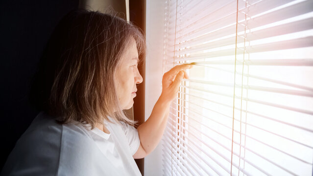 Senior woman standing by a window, peeking through horizontal blinds with her fingers, reflecting on concepts of loneliness, isolation, and hope while staying at home