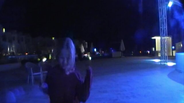A young girl seen from behind walks across a tiled resort dance floor toward bright blue spotlights in the evening with empty chairs nearby, captured on vintage analog video