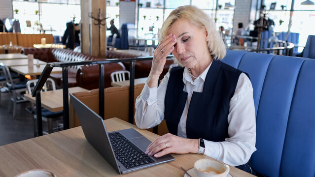 Stressful Female Professional Working. An Overwhelmed Senior Businesswoman Facing Deadlines At Busy Cafe. Distressed Professional Woman Struggles With Tasks Amidst Hurried Coffee Shop Environment