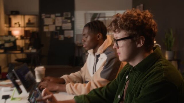 Zoom out shot of diverse young developers coding on laptops, Caucasian programmer using touchscreen tablet while African American colleague typing in office. IT startup and software engineering promo