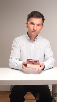 Vertical video man in a white shirt sitting at a table, holding a holy bible with a wooden cross on top, connecting with faith, spirituality, and christian religion