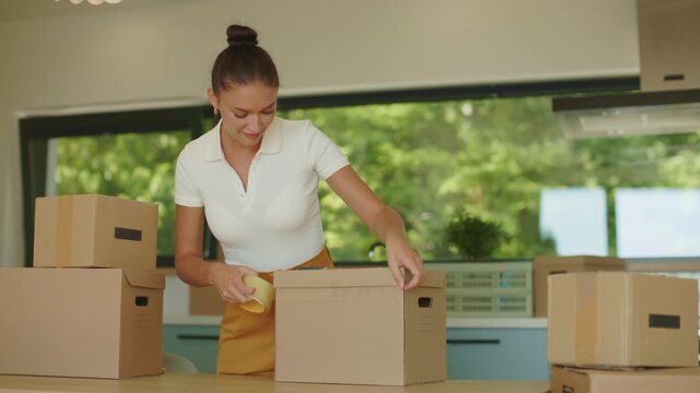 Woman sealing cardboard moving box with packing tape in bright kitchen, 4K