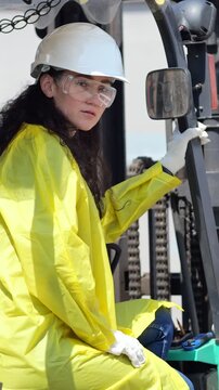 Woman In Yellow Jacket Inspecting Forklift Reprocessing Site, Operator Checking Controls With Helmet, Goggles And Gloves, Industrial Chains And Machinery In Background, Focused Contemplative