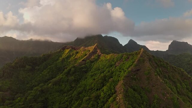 Lush green volcanic mountain ridges rise under an epic cloudy sky, showcasing the dramatic tropical landscape and natural beauty of Moorea in French Polynesia at sunset. Aerial view drone flight